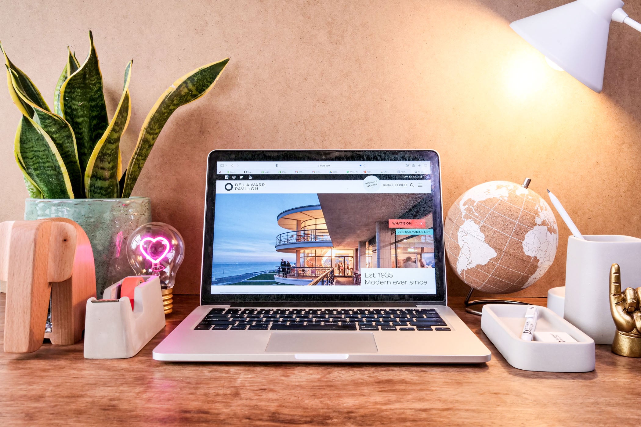 Desktop with products available from the De La Warr Pavilion online and onsite store. The image includes a laptop showing the DLWP.com website and items include a desk tidy, stapler, decorative items, a plant and a lamp