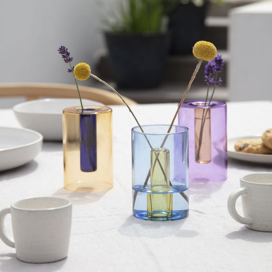 three reversible coloured glass vases with flower stems on a table set with coffee mugs
