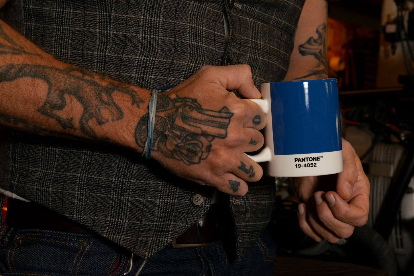 Tattooed man holding Pantone mug, blue with white handle and base. Text on white lower part reads 'PANTONE TM 19-4052'