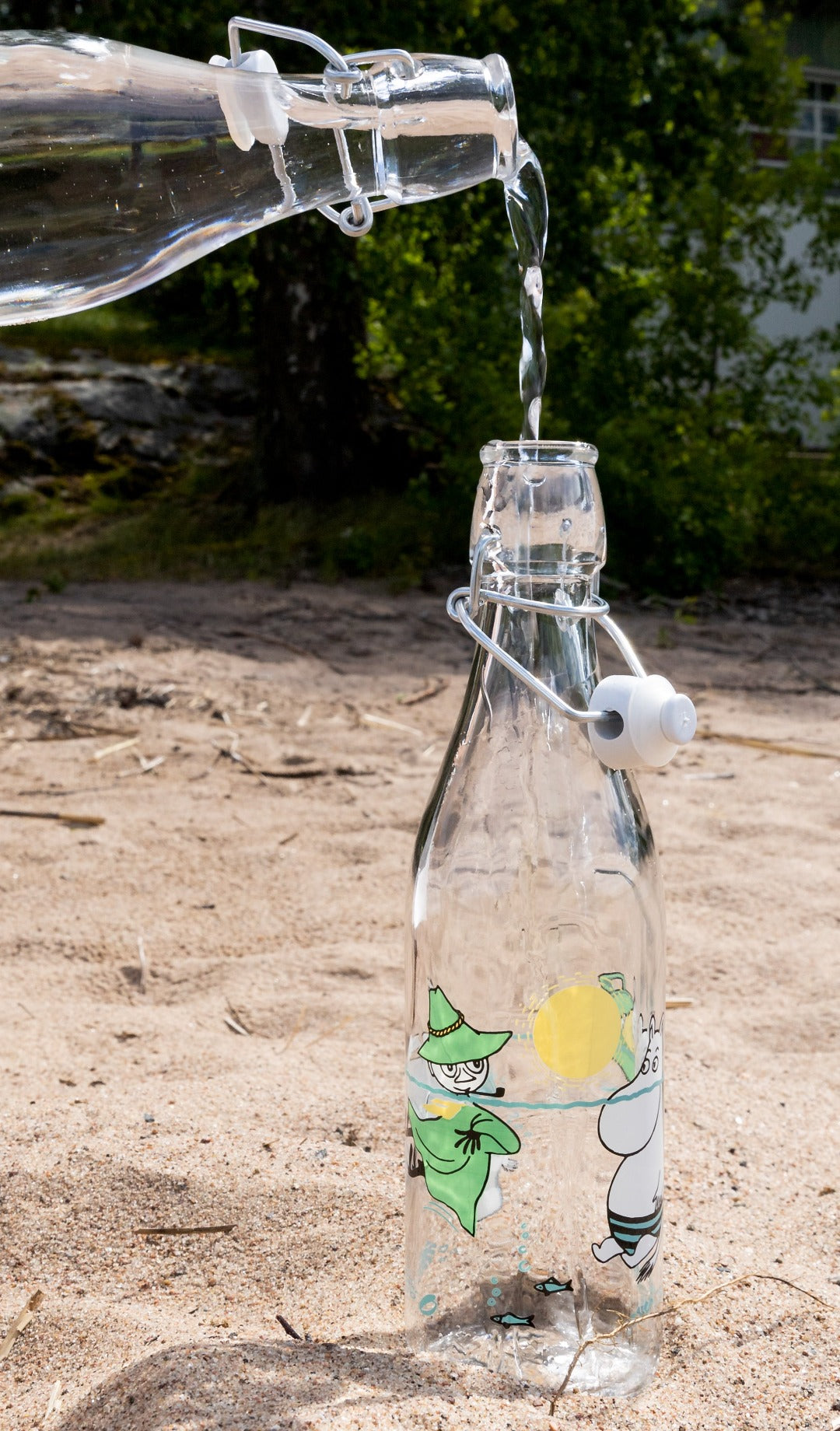 Glass bottle featuring images of Moomin characters swimming being filled on the sand