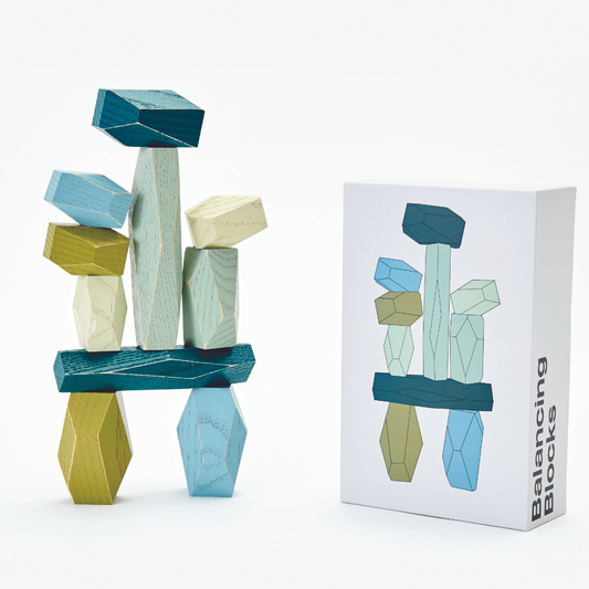 Faceted wooden shapes in various shades of blue and green, balanced on top of each other. Next to the box they come in with an illustrated image of product.