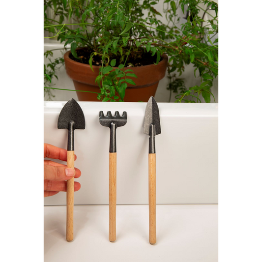 Mini rake, spade, and shovel with wooden handles. There is a potted plant in the background.