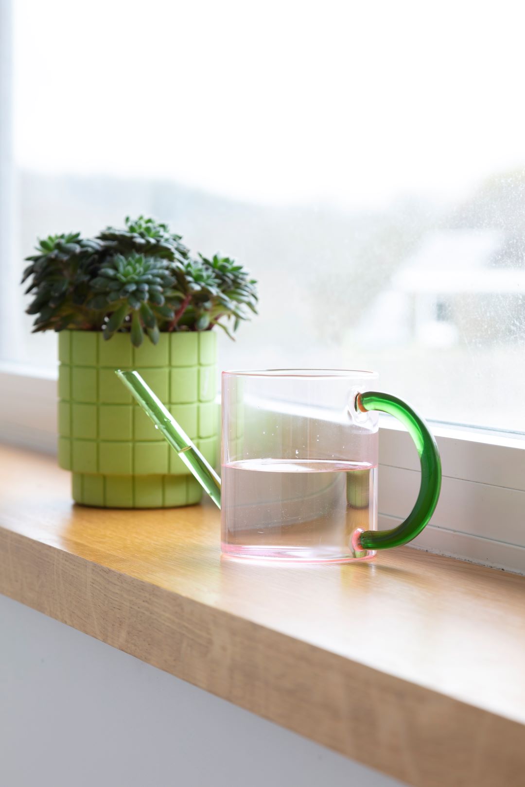 a pink glass watering can with a green handle and spout on a window sill next to a potted plant