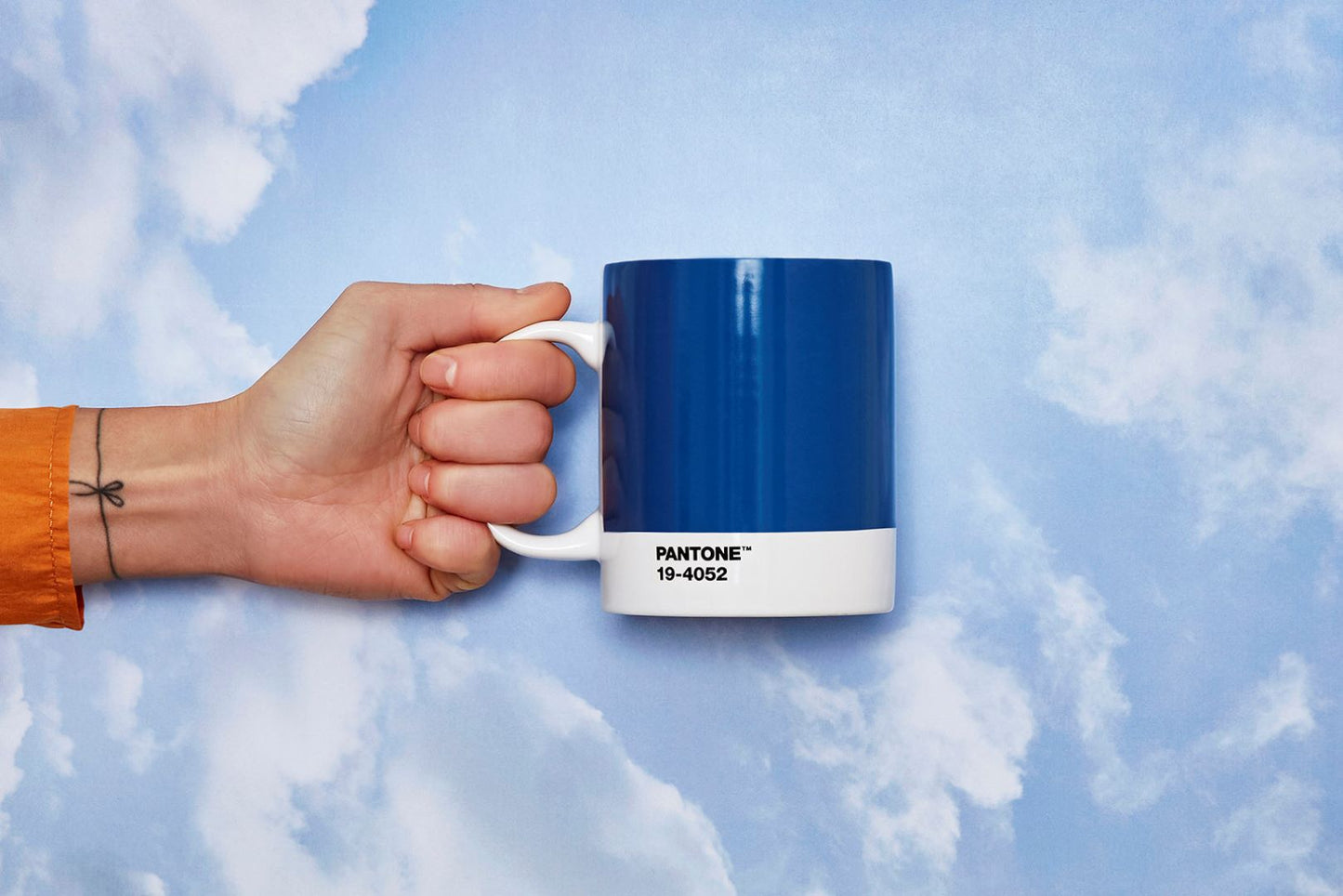 Woman holding Pantone mug, blue with white handle and base. Text on white lower part reads 'PANTONE TM 19-4052' against sky background