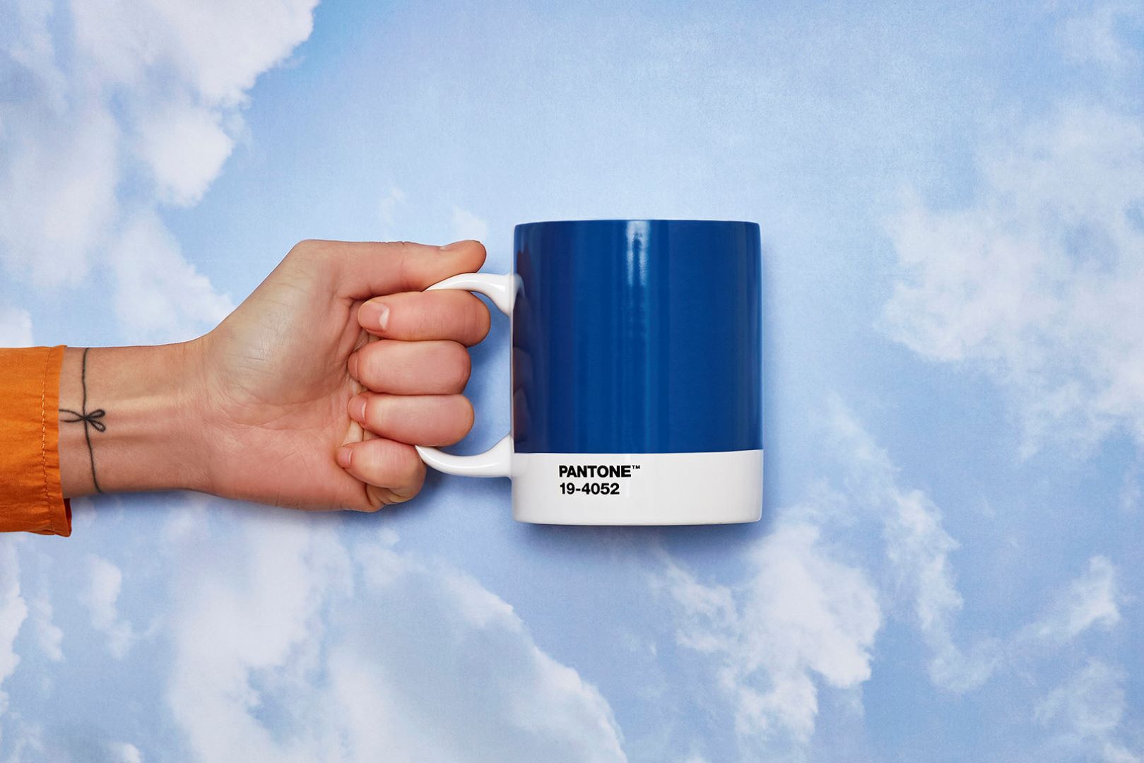 Woman holding Pantone mug, blue with white handle and base. Text on white lower part reads 'PANTONE TM 19-4052' against sky background