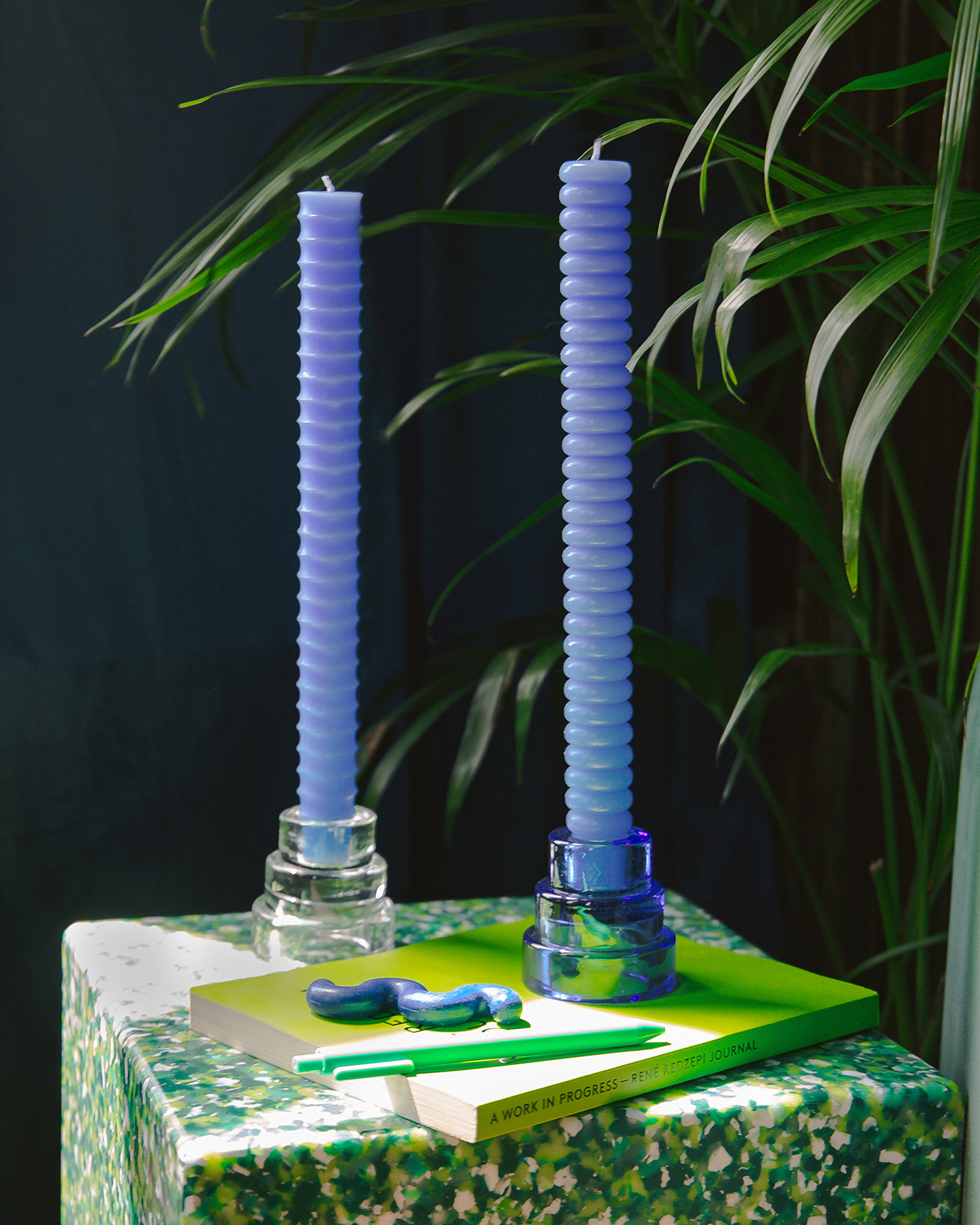 Two blue bumpy taper candles in a clear standon a green table with a book.