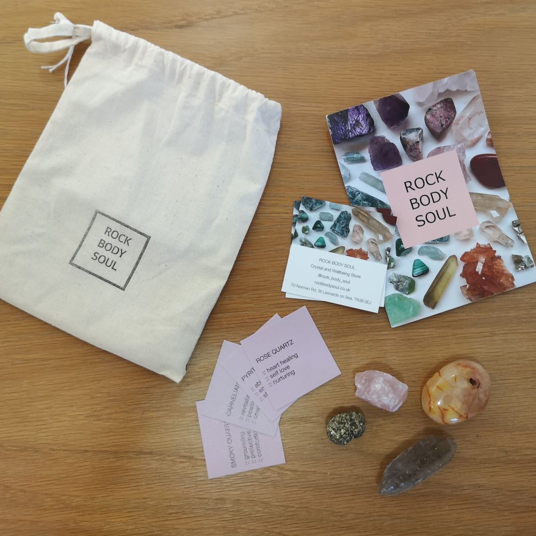rose quartz, pyrite, carnelian, and smoky quartz on a wooden surface, with 'Rock Body Soul' info cards and white drawstring bag.