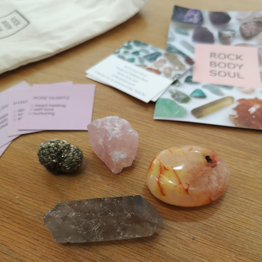 rose quartz, pyrite, carnelian, and smoky quartz on a wooden surface, with 'Rock Body Soul' info cards and white drawstring bag.