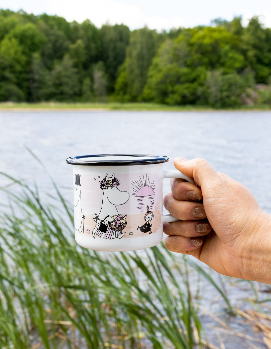 someone holding enamel Moomin mug with characters at the beach in front of lake