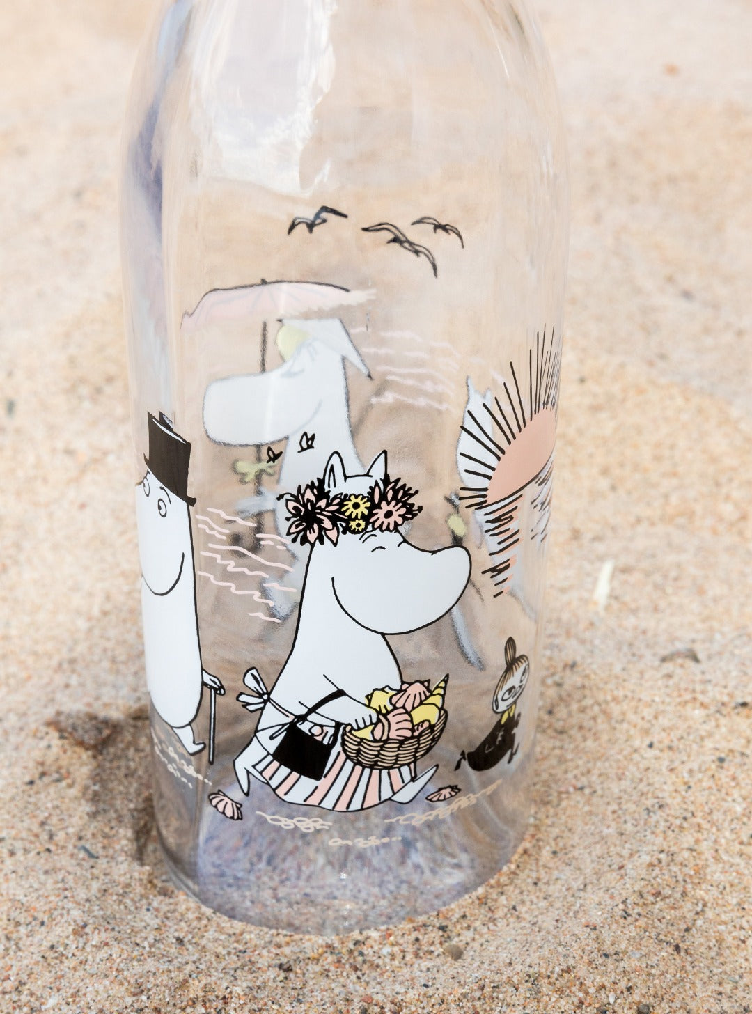 Glass bottle featuring Moomin characters at the beach, placed on the sand