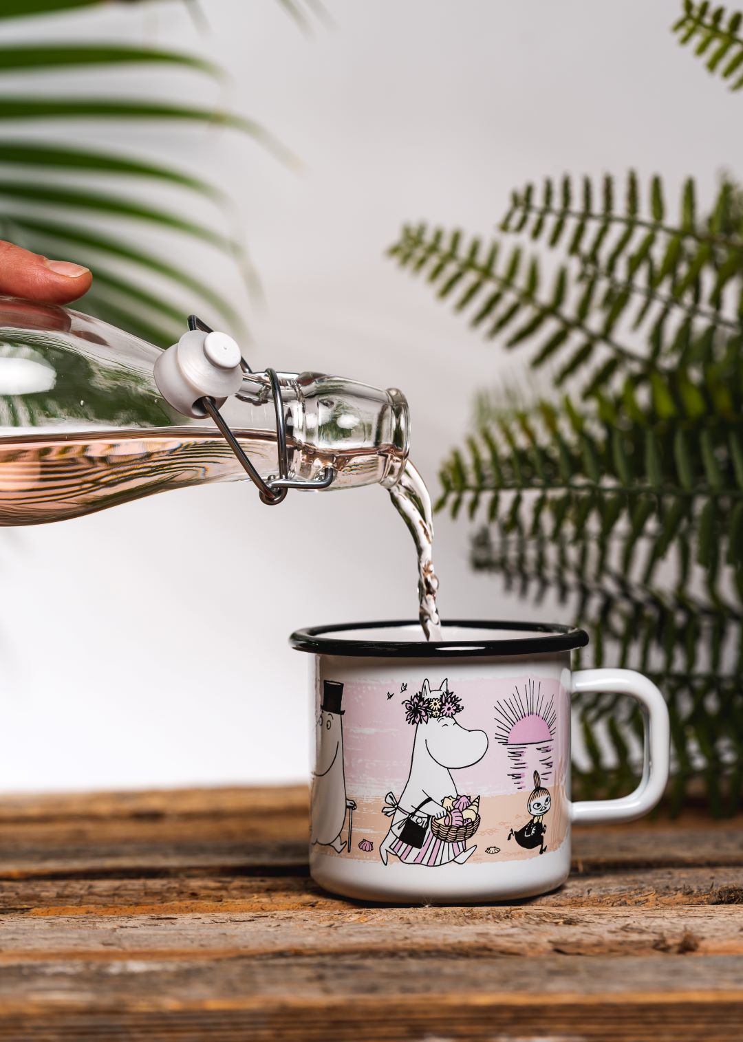 someone pouring drink into enamel Moomin mug with characters at the beach