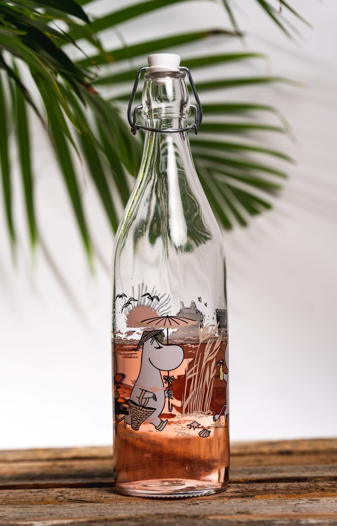 Glass bottle featuring Moomin characters at the beach, filled with a pale pink drink on a table