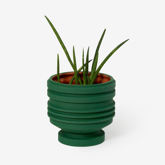 Green plant pot with sculptural ridges and shape, and a terracotta glazed inside. With a plant inside. 