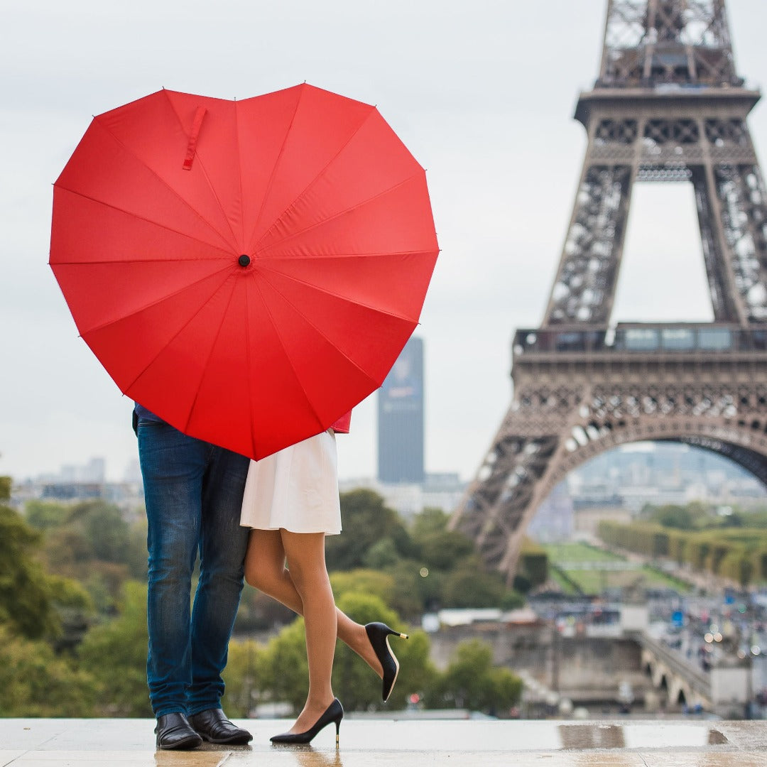 Couple under a red heart shaped umbrella, with the Eiffel Tower in the backgorund.